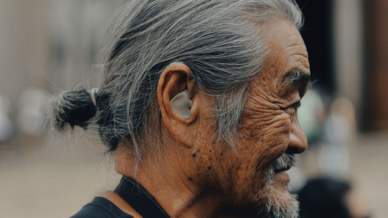 Close profile of an older man with grey hair tied back, a hearing aid visible in his ear, outdoors in soft light