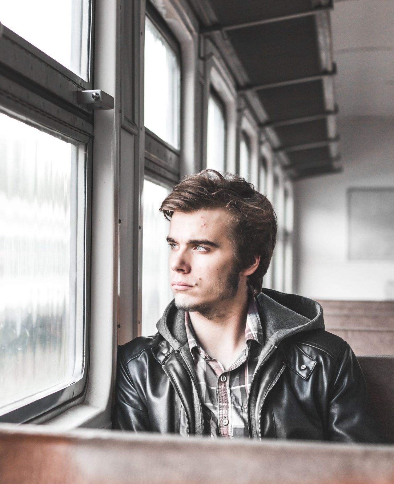 A young man in a leather jacket sitting on a train, looking out of the window