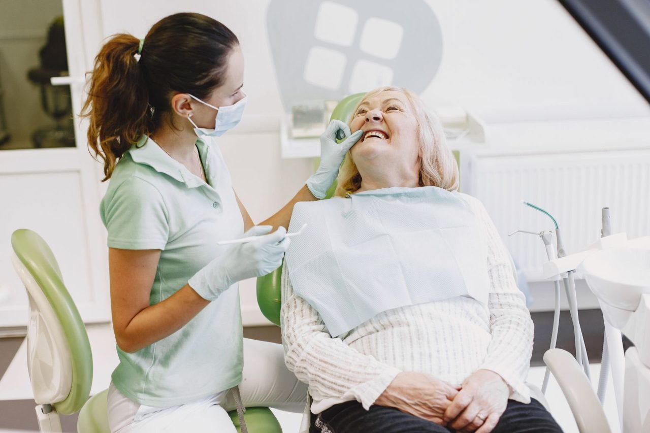 An older woman laughing in a dental chair while a dentist in a mask and gloves examines her teeth