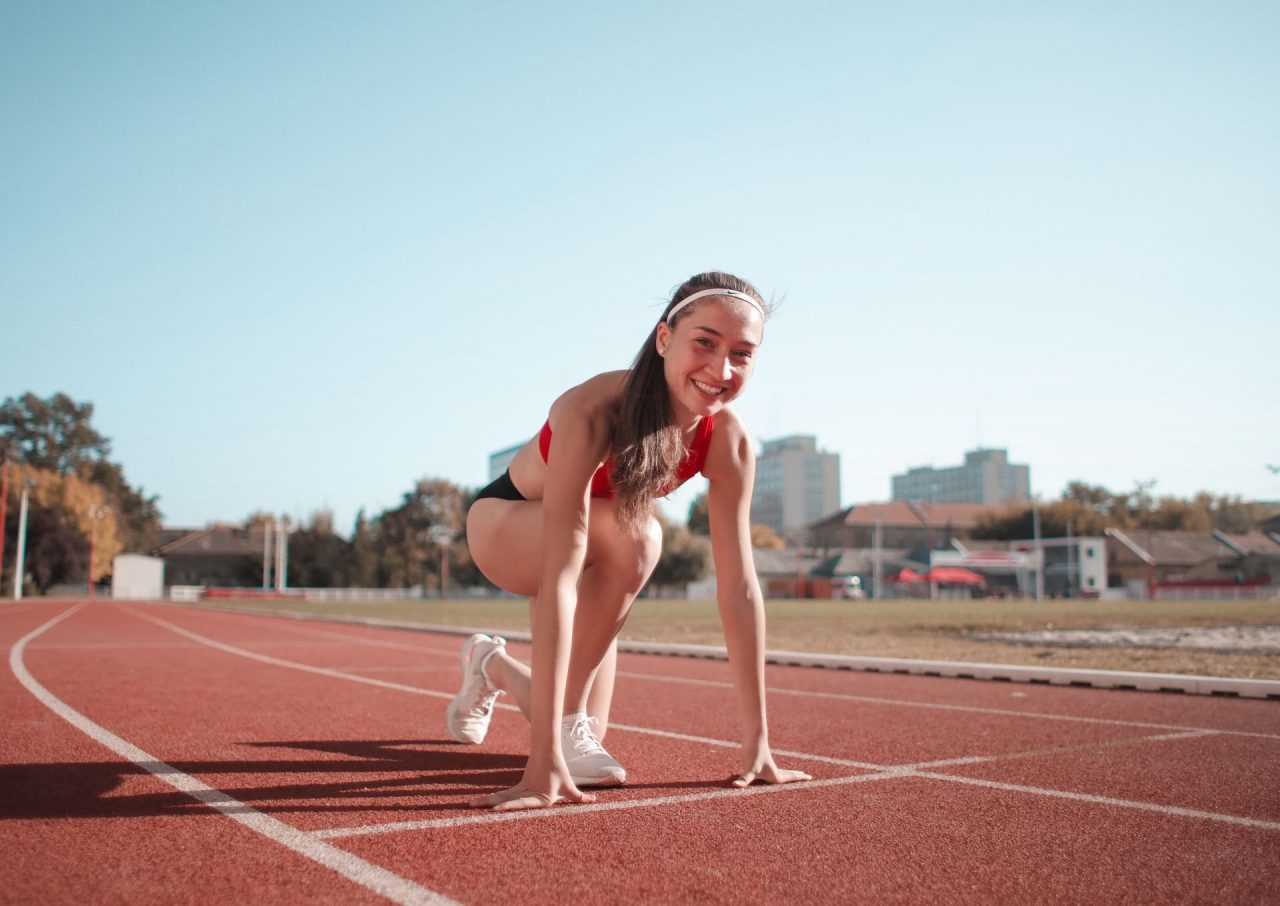 A smiling young woman crouched at the start line of an outdoor running track on a sunny day