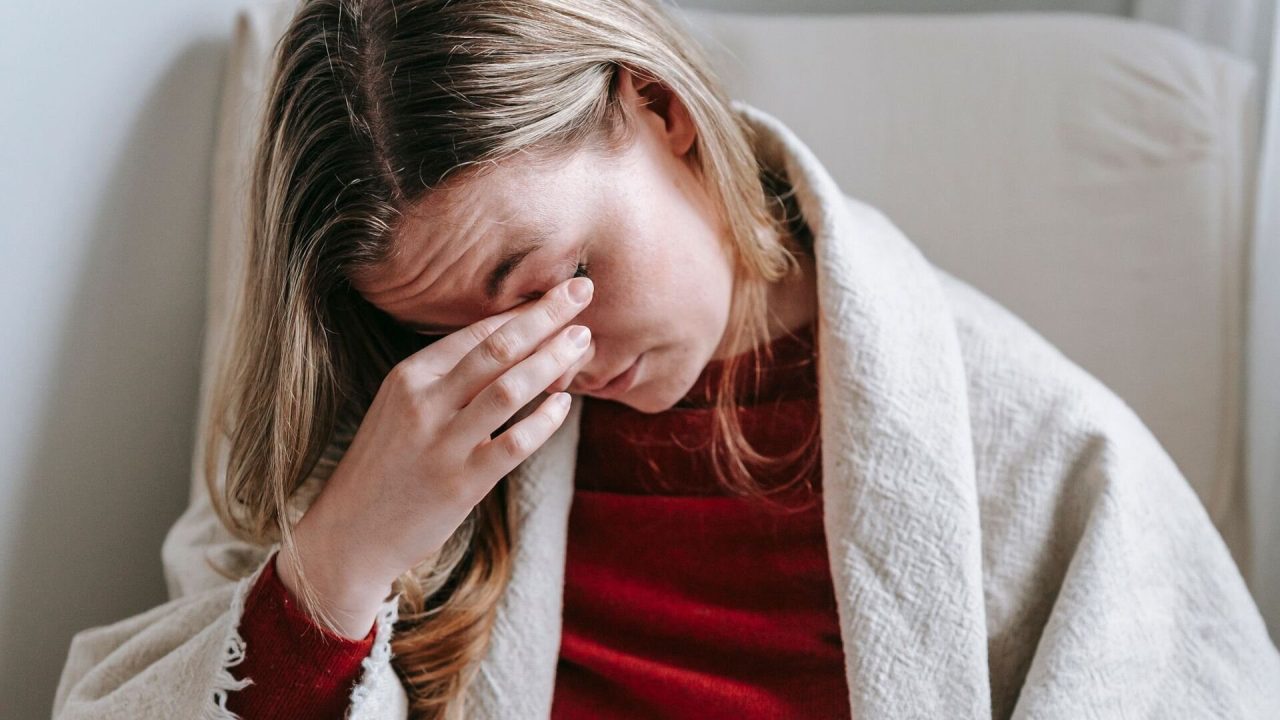 A woman wrapped in a blanket sitting in an armchair, pressing her fingers to her tired eyes