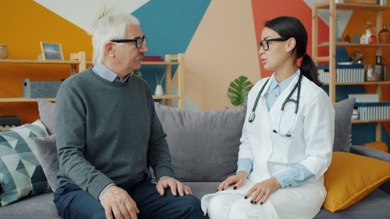 An older man in a grey jumper talking with a doctor in a white coat in a bright clinic room