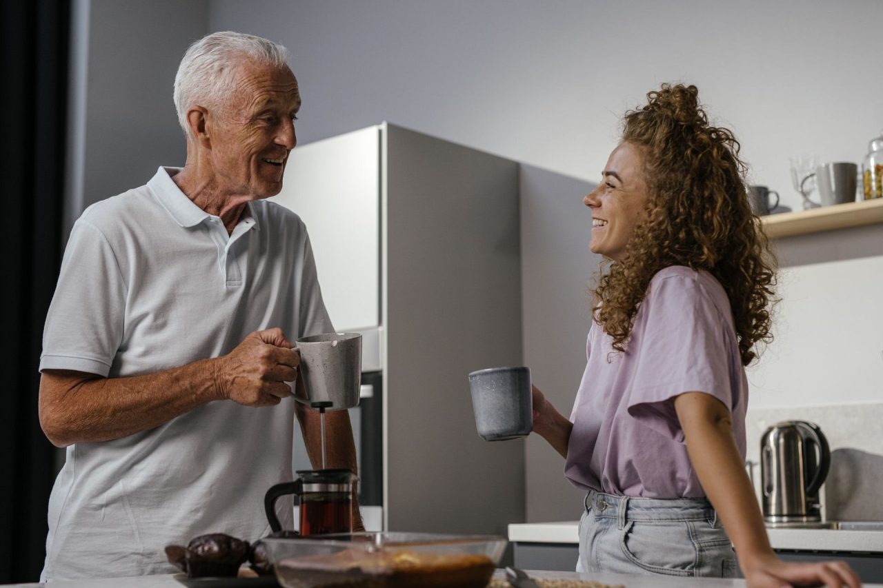 An older man and his adult granddaughter laughing together over coffee in a bright kitchen