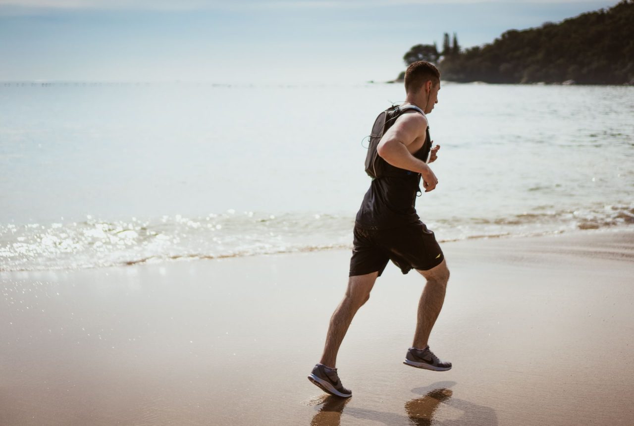 A man in a black singlet and shorts running along the wet sand at the edge of the sea
