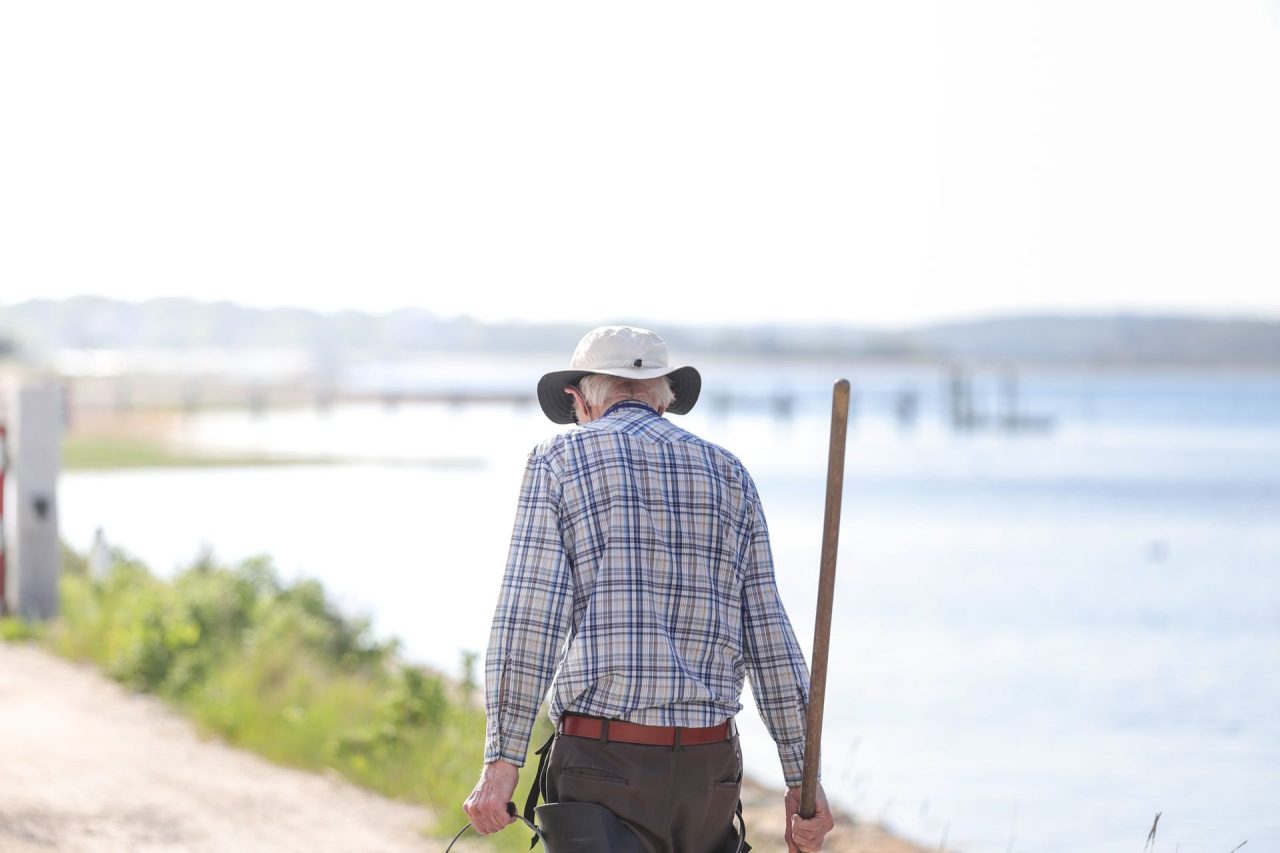 An older man in a checked shirt and sun hat walking with a stick along a waterside path on a sunny day
