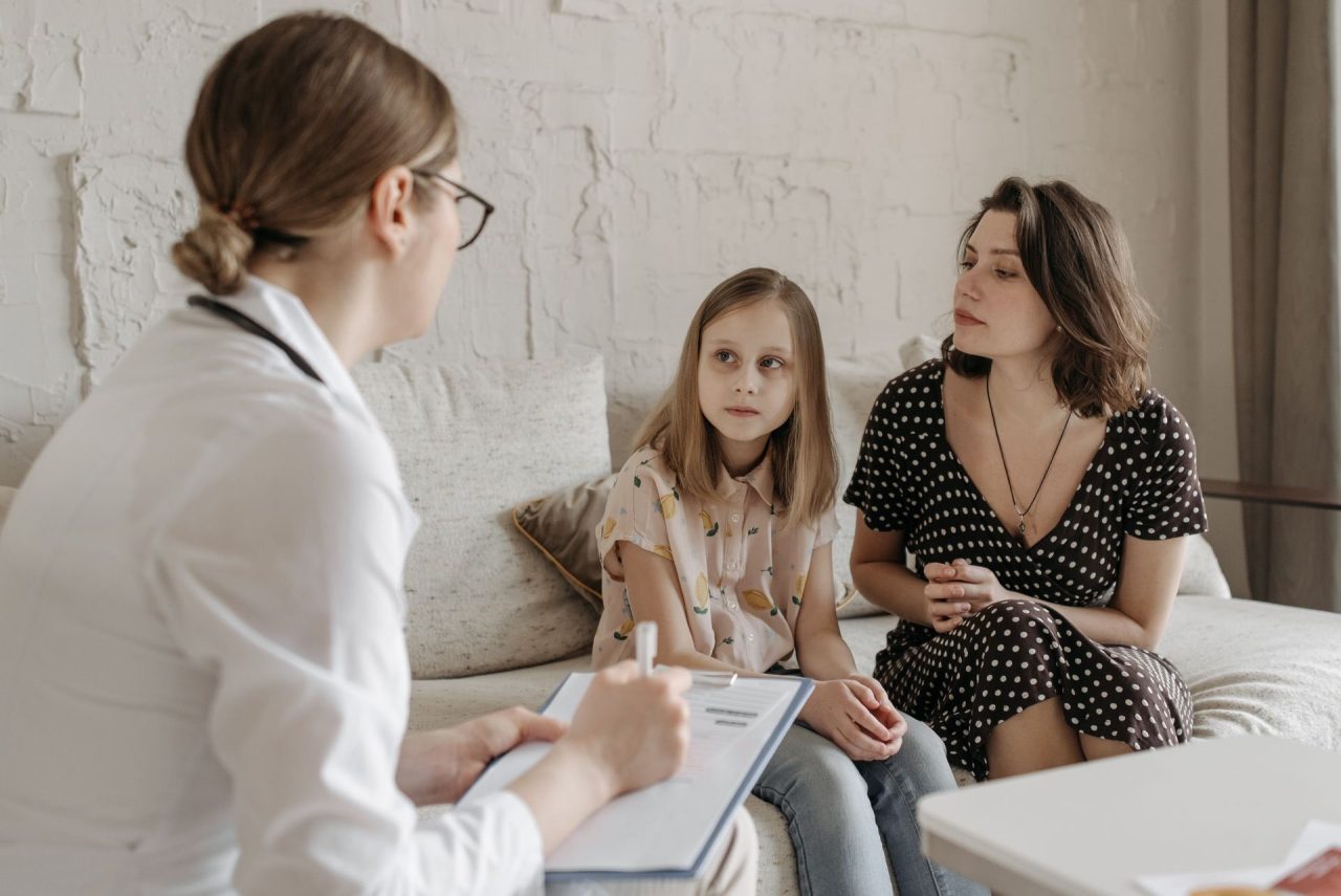 A doctor taking notes while talking with a young girl and her mother on a sofa in a bright consulting room