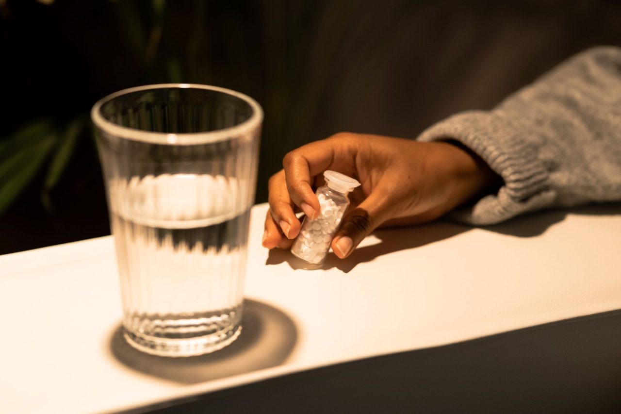 A hand reaching for a small bottle of tablets beside a full glass of water on a sunlit bench