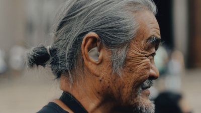 Close profile of an older man with grey hair tied back, a hearing aid visible in his ear, outdoors in soft light