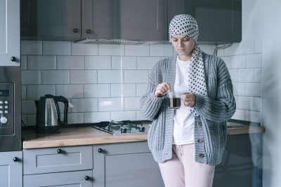 A woman in a patterned headscarf and grey cardigan holding a cup of tea in her kitchen