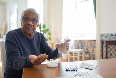 An older man sitting at his kitchen table with a glass of water and his morning tablets laid out in front of him