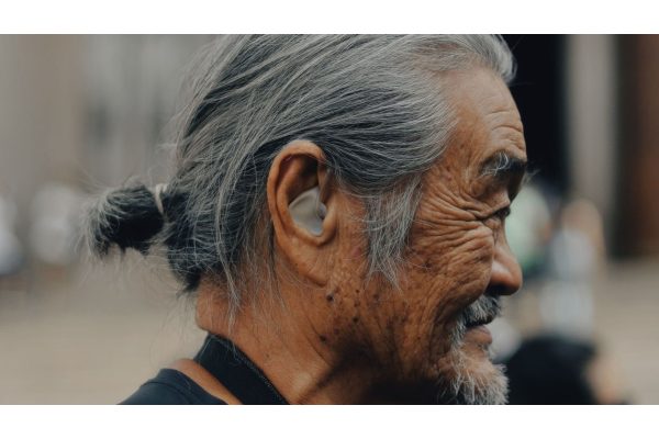 Close profile of an older man with grey hair tied back, a hearing aid visible in his ear, outdoors in soft light