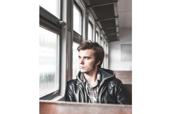 A young man in a leather jacket sitting on a train, looking out of the window