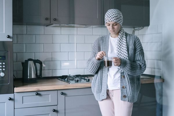 A woman in a patterned headscarf and grey cardigan holding a cup of tea in her kitchen