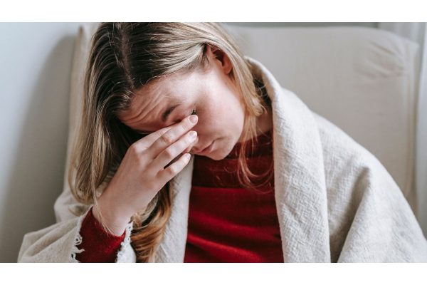 A woman wrapped in a blanket sitting in an armchair, pressing her fingers to her tired eyes