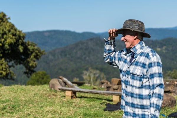 A farmer in a checked shirt and hat shading his eyes as he looks across hills on a clear day