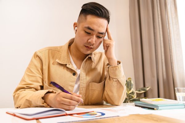 A young man in a tan jacket pausing with his hand to his temple while writing notes at a desk