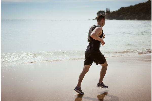 A man in a black singlet and shorts running along the wet sand at the edge of the sea