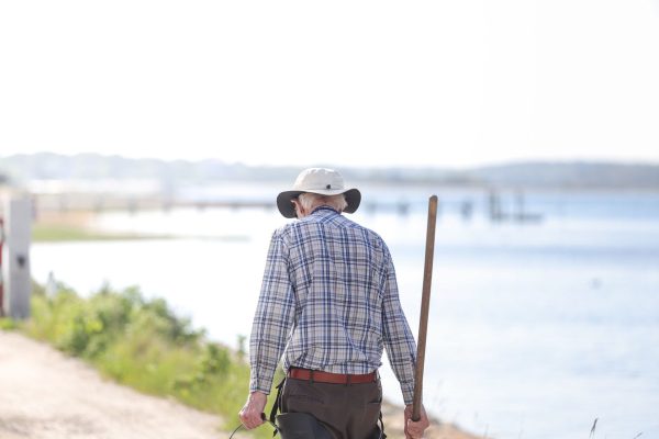An older man in a checked shirt and sun hat walking with a stick along a waterside path on a sunny day
