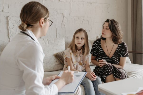 A doctor taking notes while talking with a young girl and her mother on a sofa in a bright consulting room