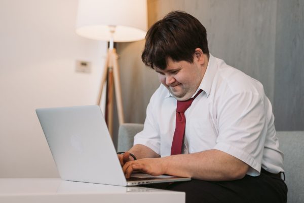 A smiling young man with Down syndrome in a white shirt and red tie working on a laptop at home