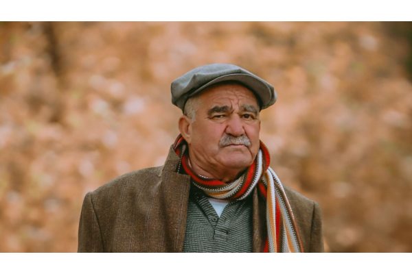 An older man in a flat cap and tweed jacket with a striped scarf around his neck, standing outdoors in autumn